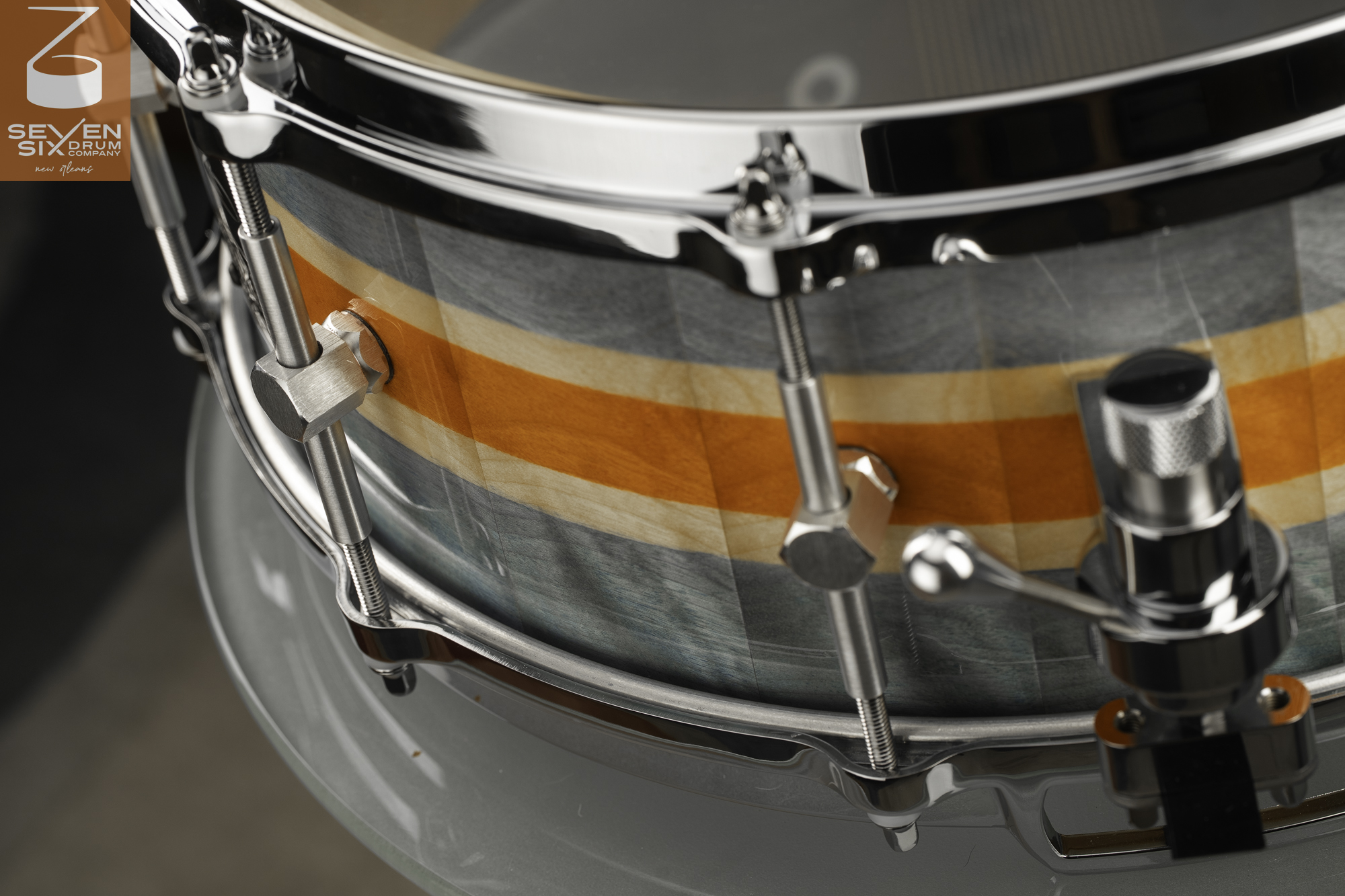 Handmade snare drum featuring an orange and gray striped wood shell with polished chrome hoops, lugs, and a throw-off.