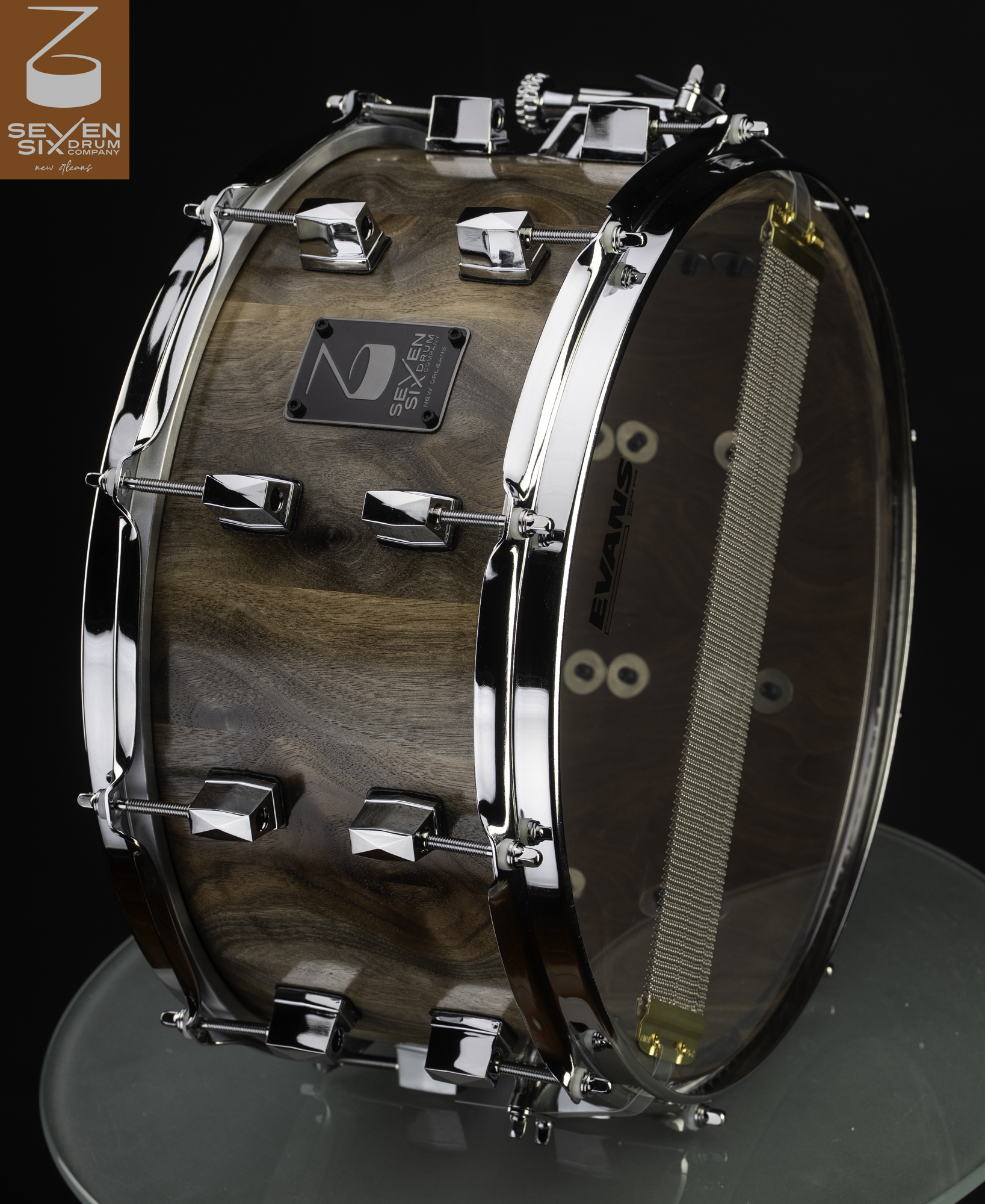 Natural satin walnut snare drum with polished chrome lugs, hoops, and a clear bottom head.