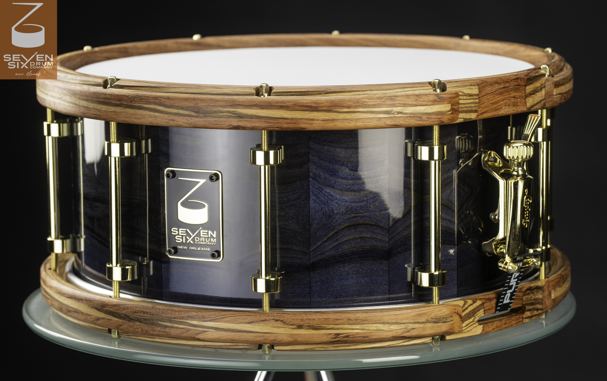 Handmade snare drum with a glossy blue wood grain shell, striped wooden hoops, and polished brass tube lugs.