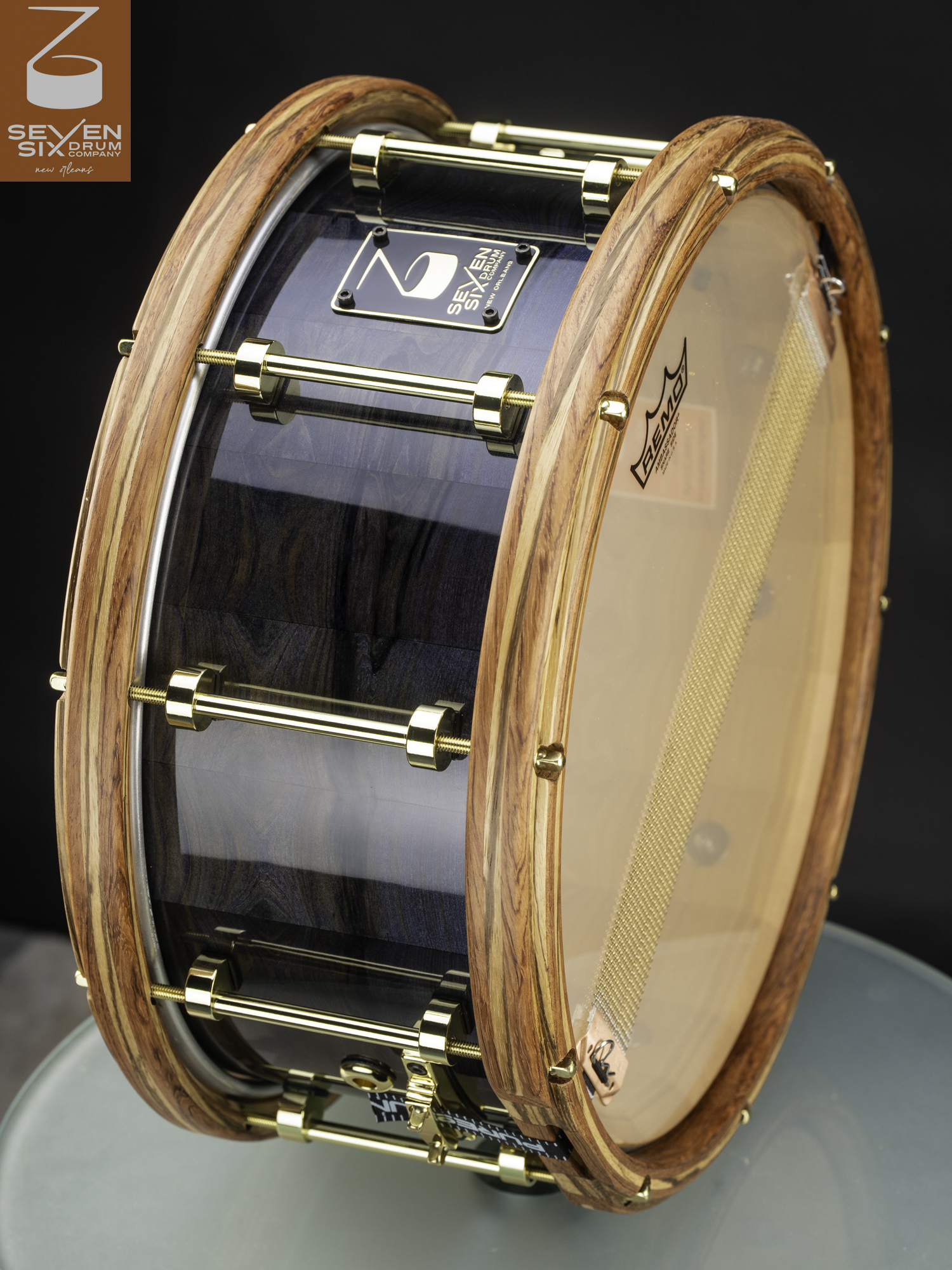 Handmade snare drum with a glossy blue-gray wood shell, natural wood hoops, and polished brass tube lugs.