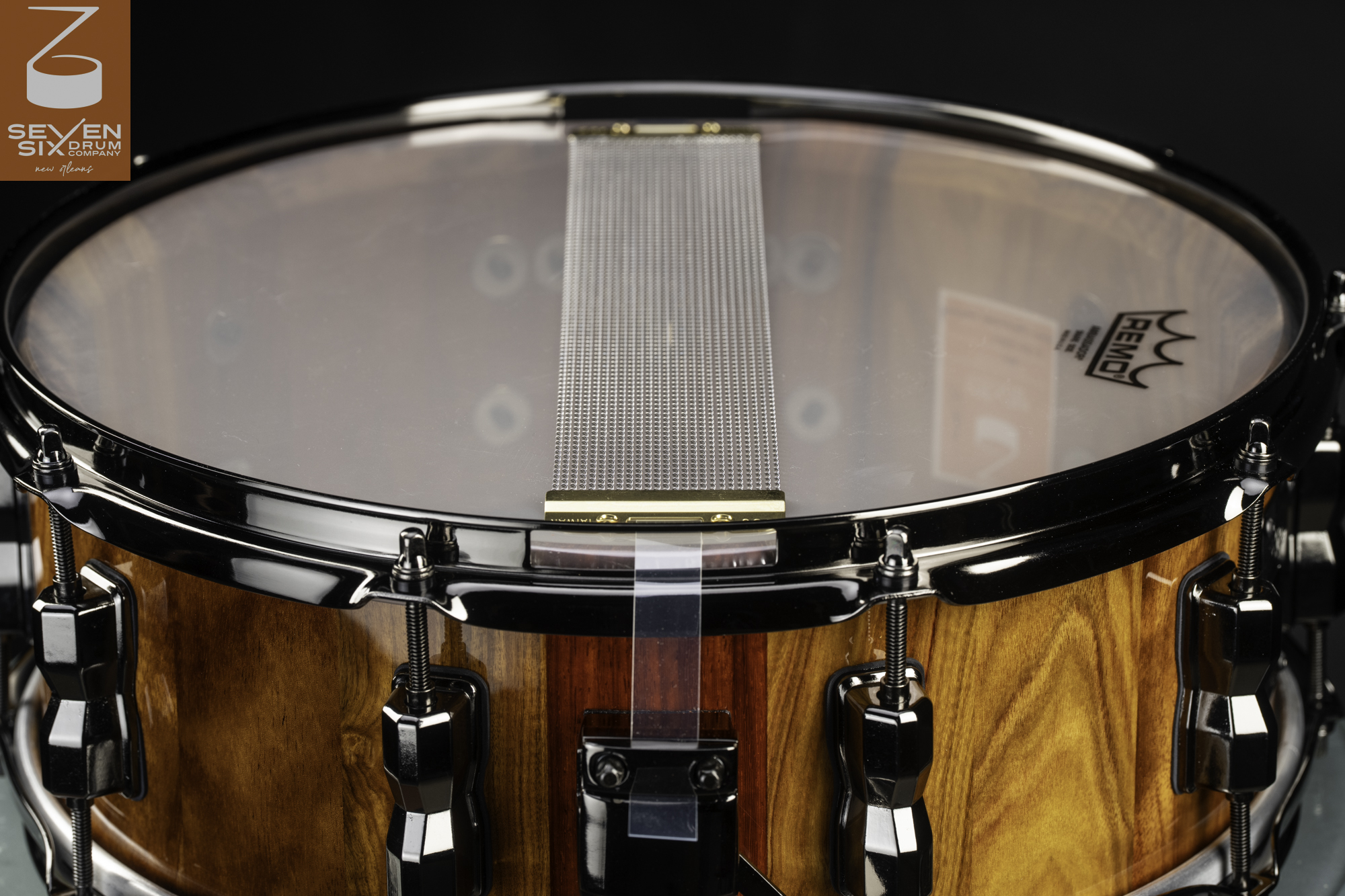 Handmade snare drum with a glossy natural wood shell, black nickel hardware, and wide metal snare wires over a clear head.