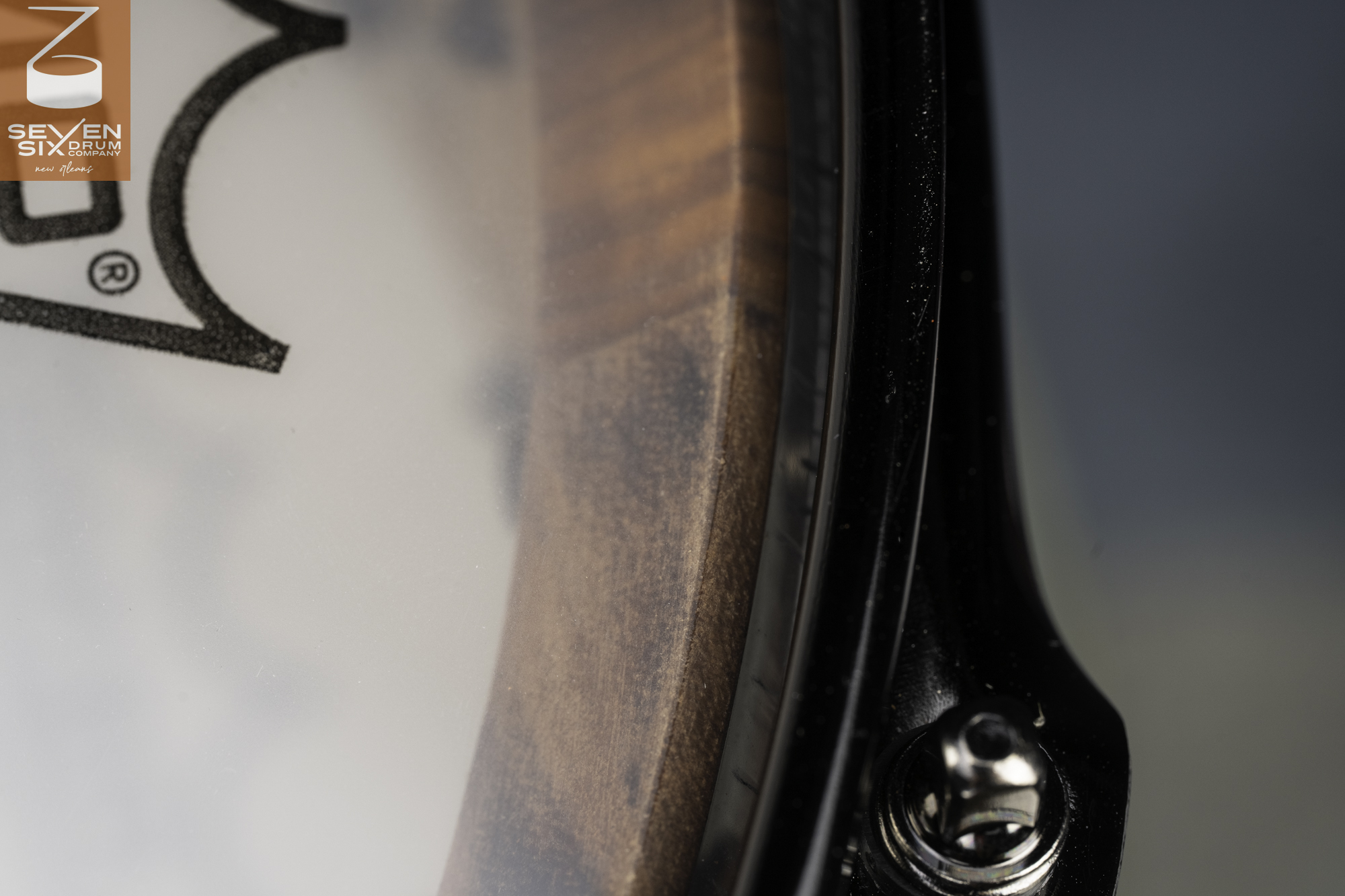 Close-up of a dark natural wood snare drum shell with a black metal hoop and chrome tension rod.