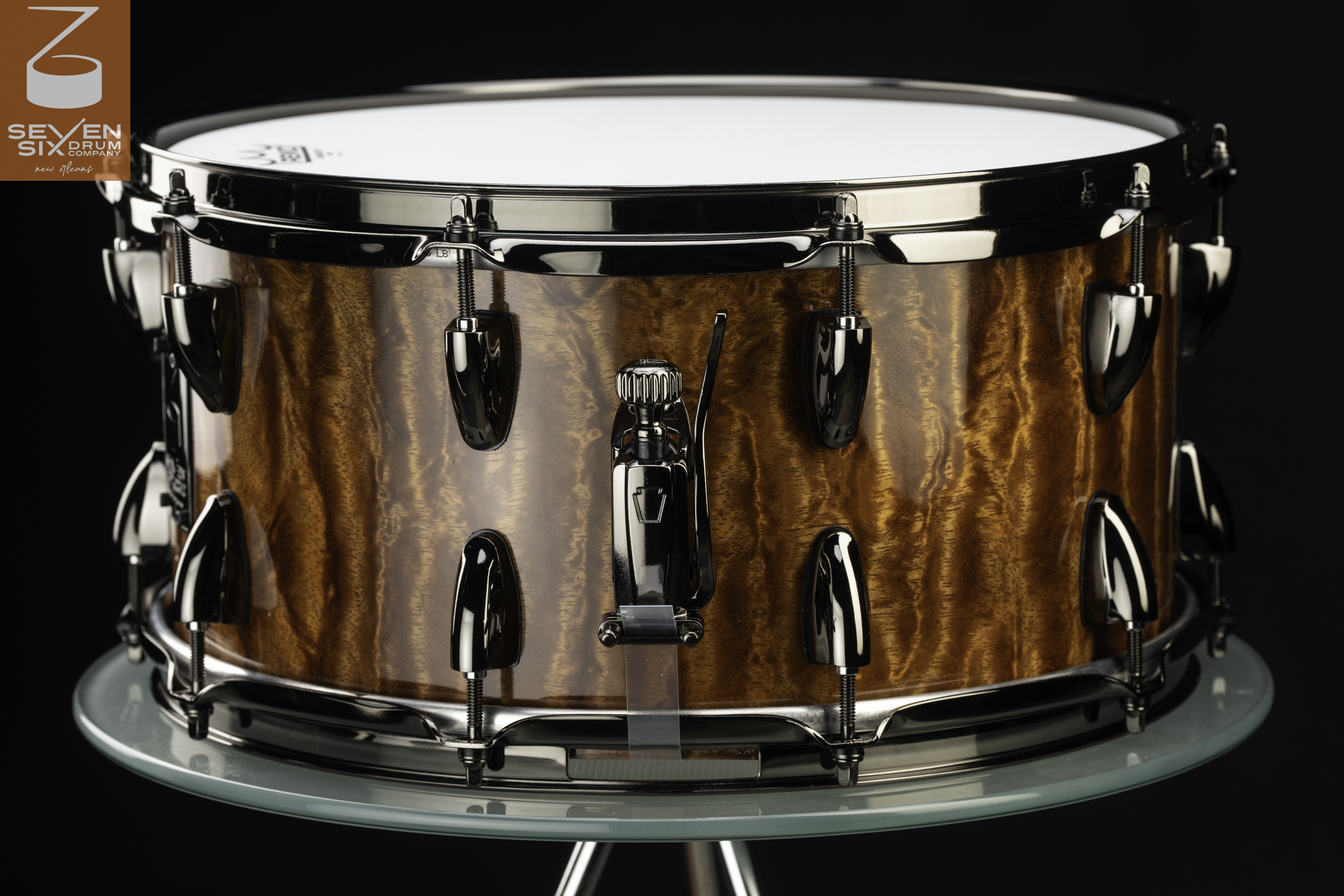 Handmade snare drum with a glossy figured wood shell, black nickel hoops, teardrop lugs, and a matching throw-off.