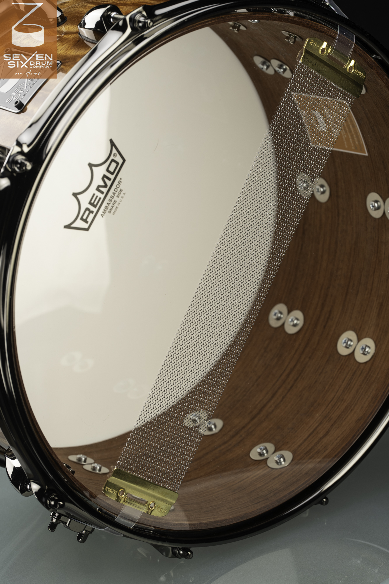 Underside of a wooden snare drum featuring a clear Remo head, wide wire snare with brass clips, and dark chrome hardware.