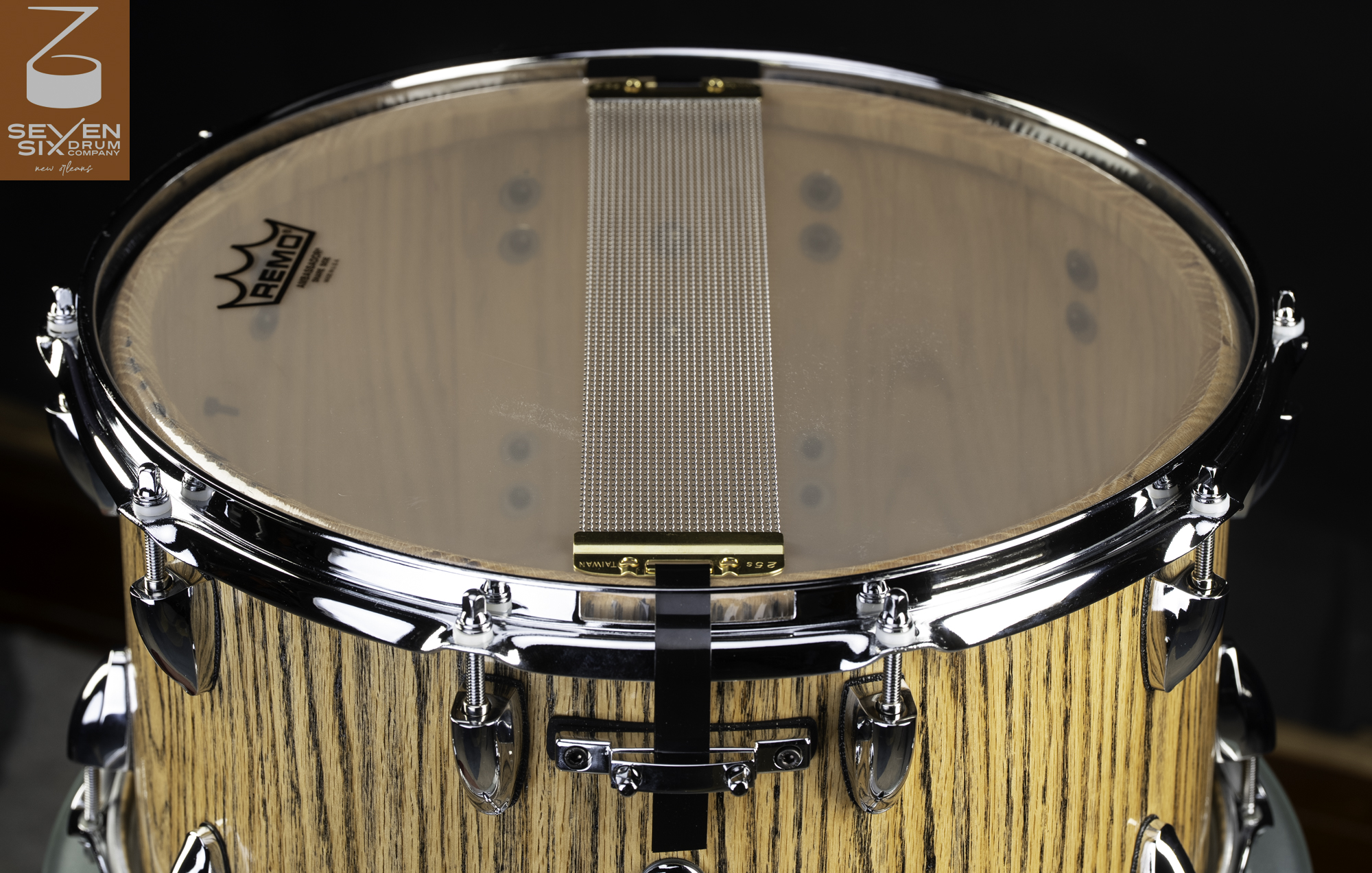 Bottom view of a natural wood snare drum showing chrome hardware and snare wires mounted across a clear drumhead.