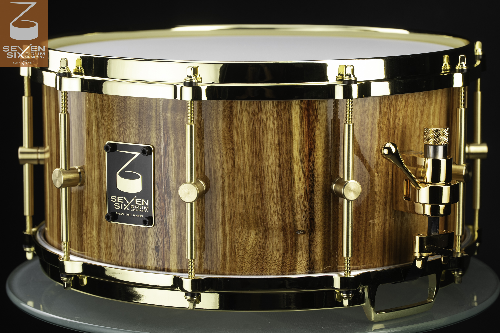 Handcrafted wooden snare drum with a glossy natural grain shell, polished brass hoops, tube lugs, and gold throw-off.