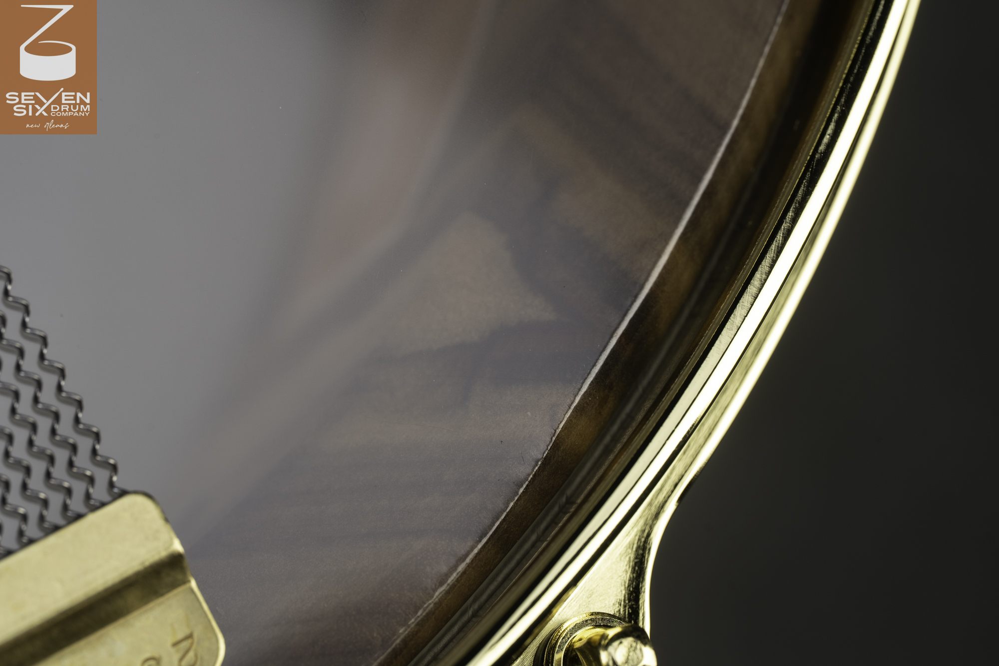 Close-up of a snare drum with a clear head showing a glossy dark wood shell, polished brass hoop, and snare wires.