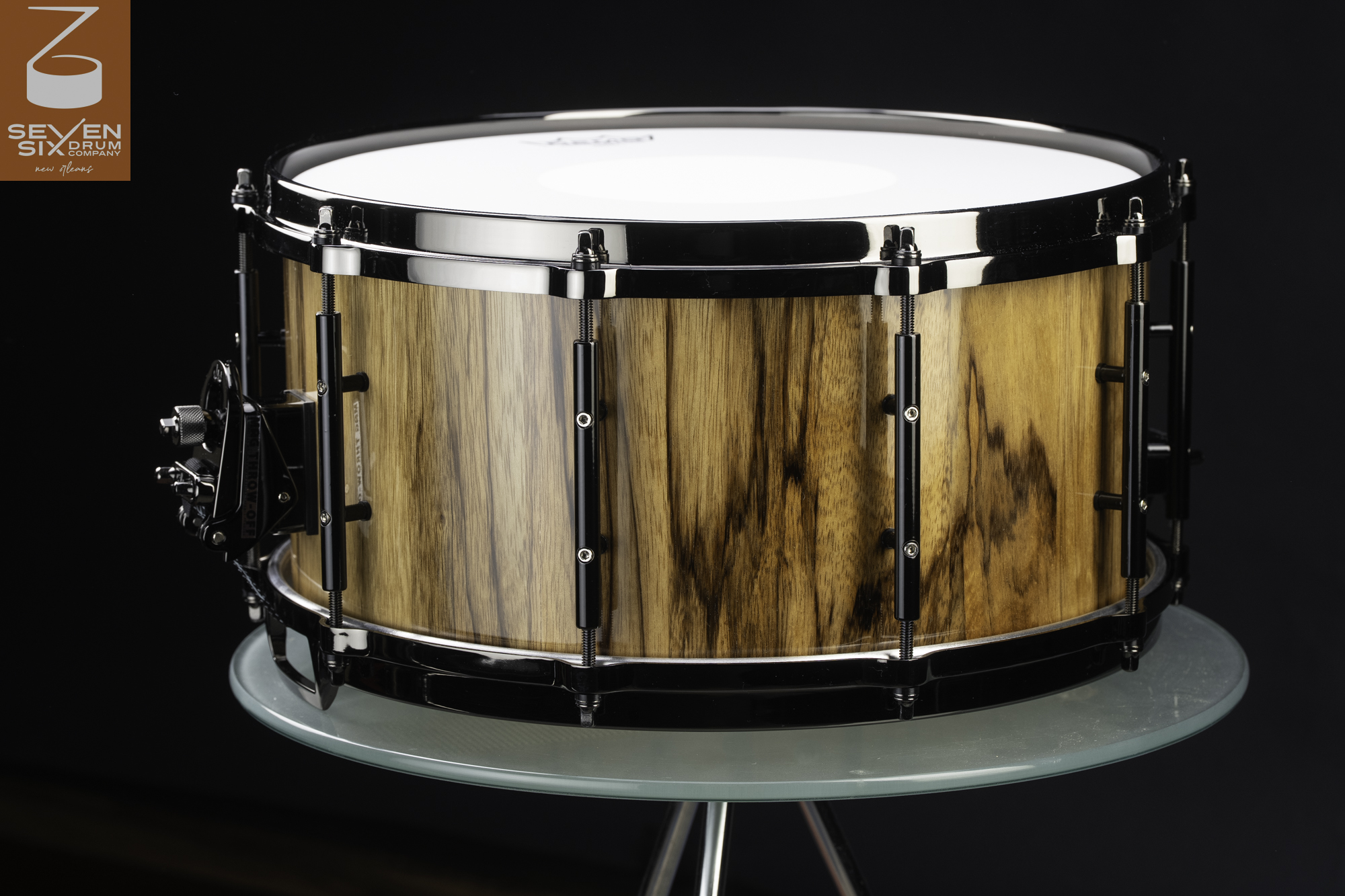 Handmade snare drum with a gloss-finished figured wood shell, black nickel hoops, tube lugs, and a throw-off mechanism.