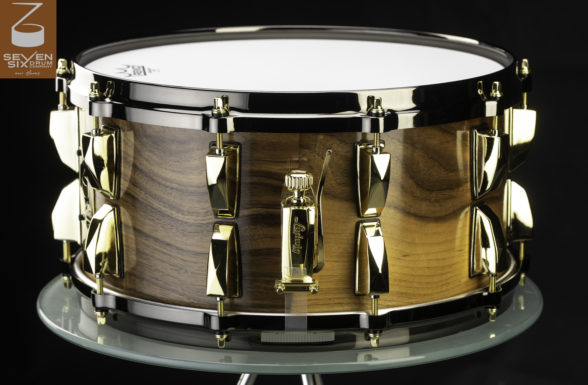 Handmade snare drum featuring a gloss wood shell, black nickel hoops, and polished gold lugs with a matching throw-off.