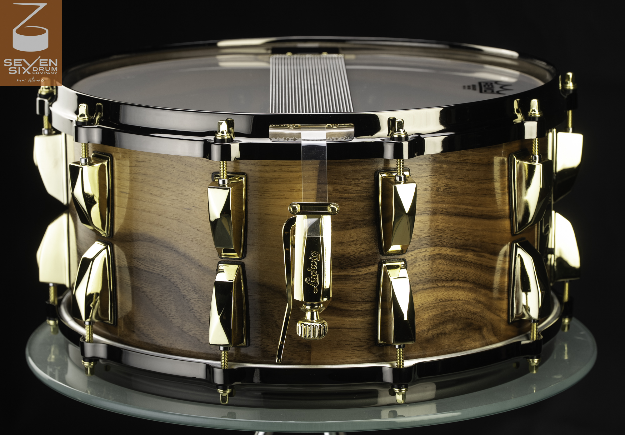 Handmade wooden snare drum with a glossy natural finish, gold lugs and strainer, and dark chrome hoops.
