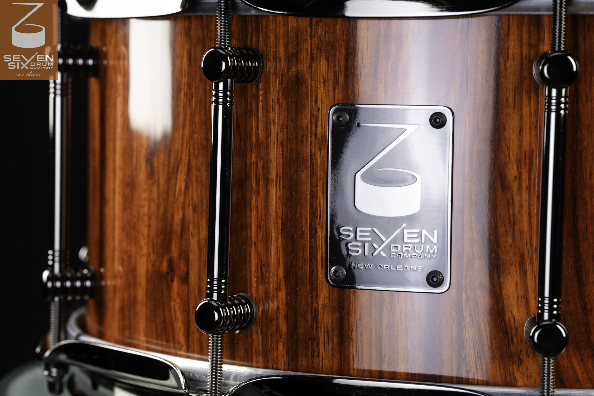 Handmade snare drum with a glossy reddish-brown wood finish, black chrome tube lugs, and a polished metal badge.