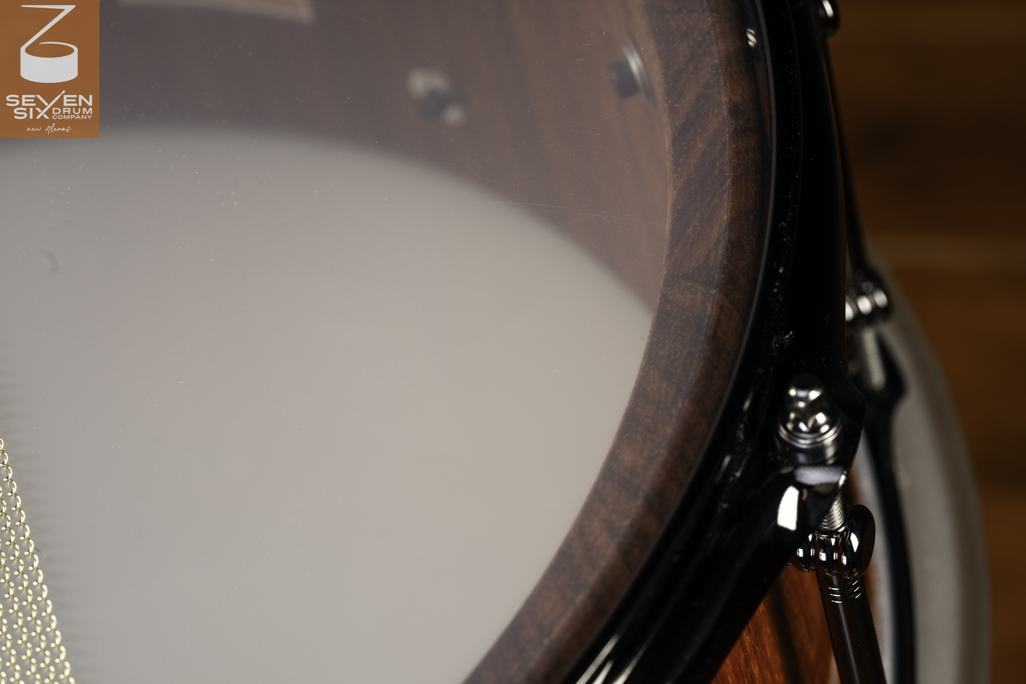 Close-up of a wooden snare drum with a rich brown finish, clear resonant head, black hoop, and chrome tension rod.