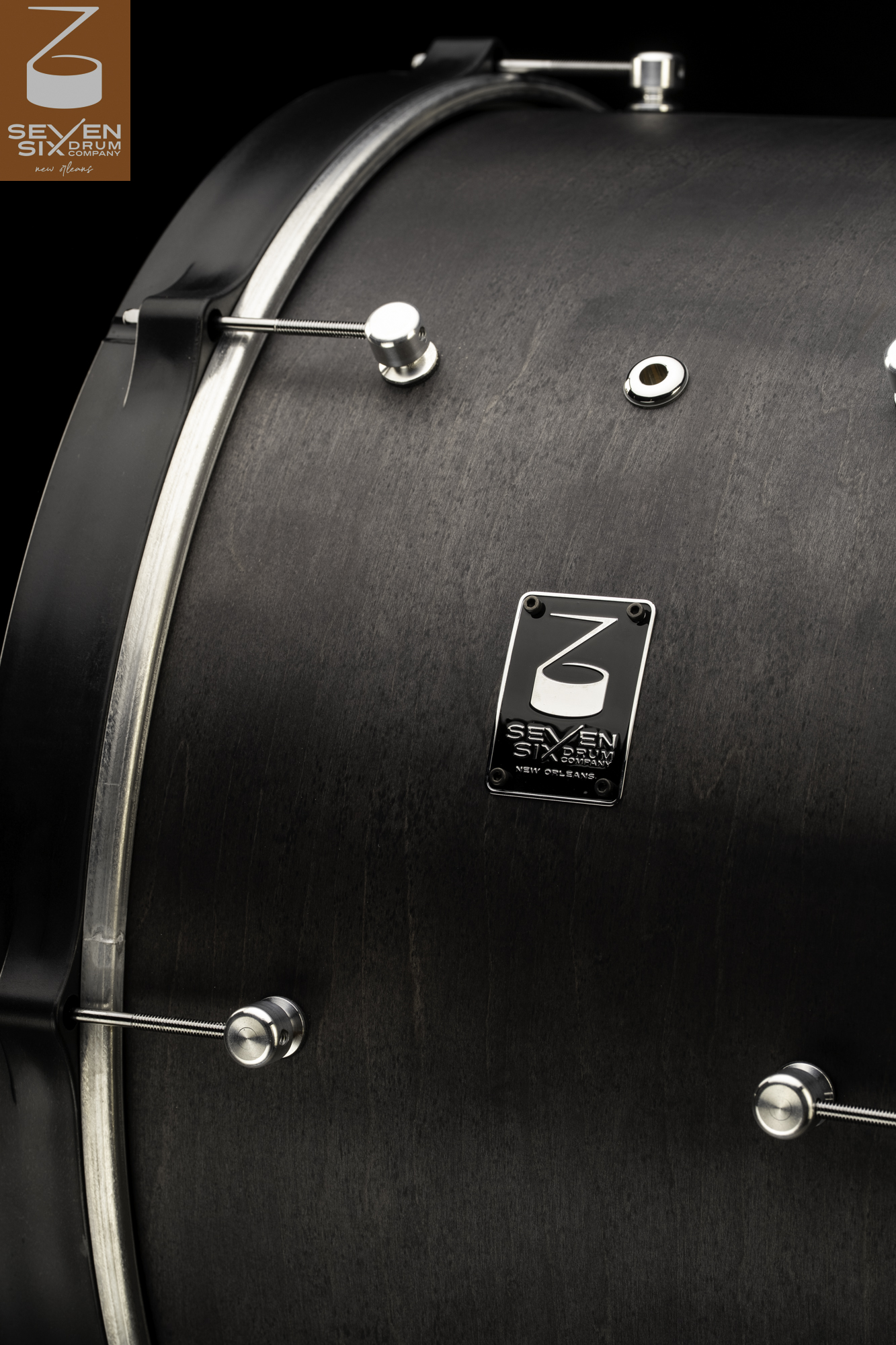 Dark charcoal stained wood drum featuring minimalist chrome lugs, a black hoop, and a branded metal badge.