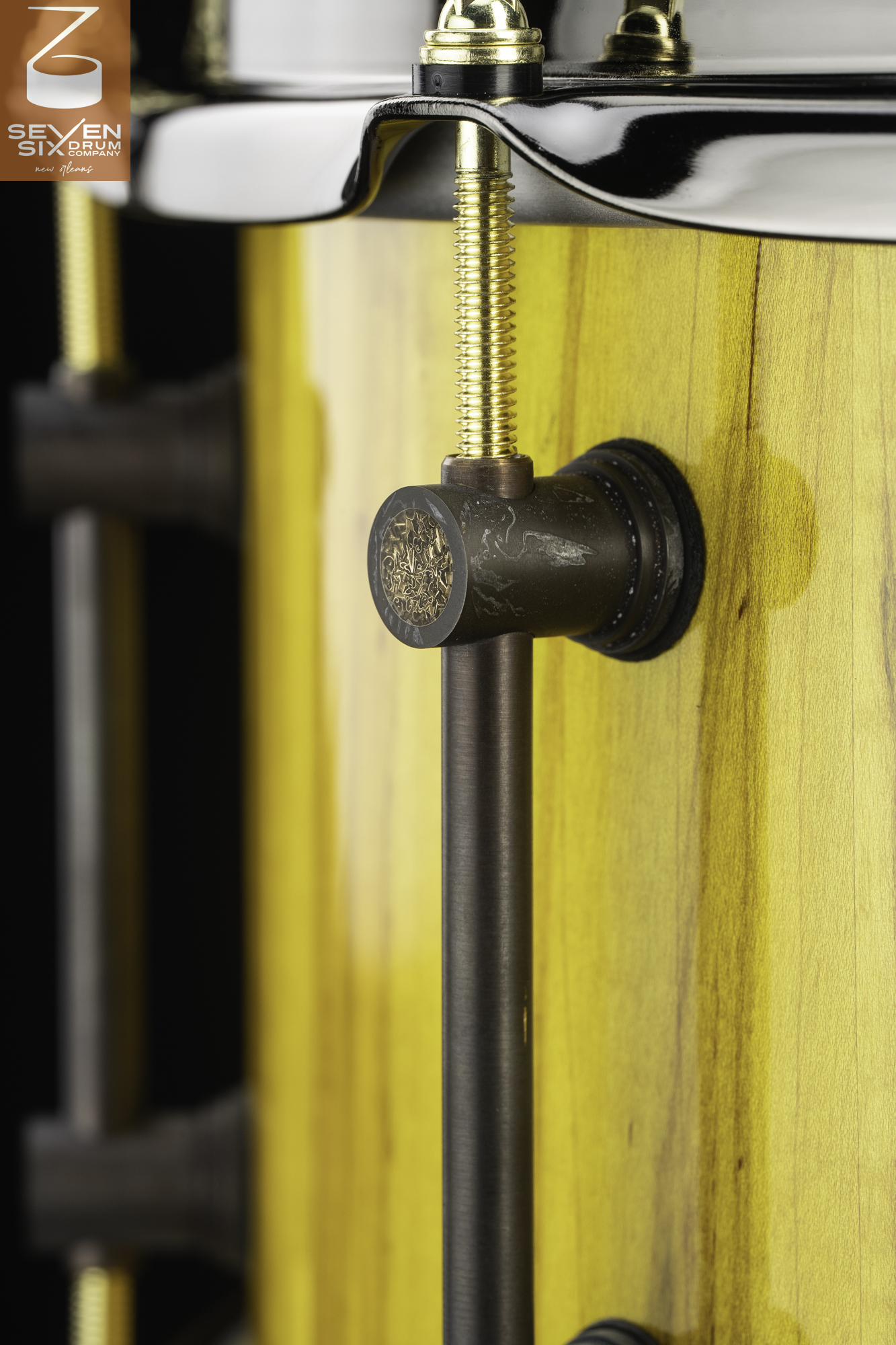 Detail of a glossy yellow wood snare drum with an antique bronze tube lug, gold tension rod, and black hoop.