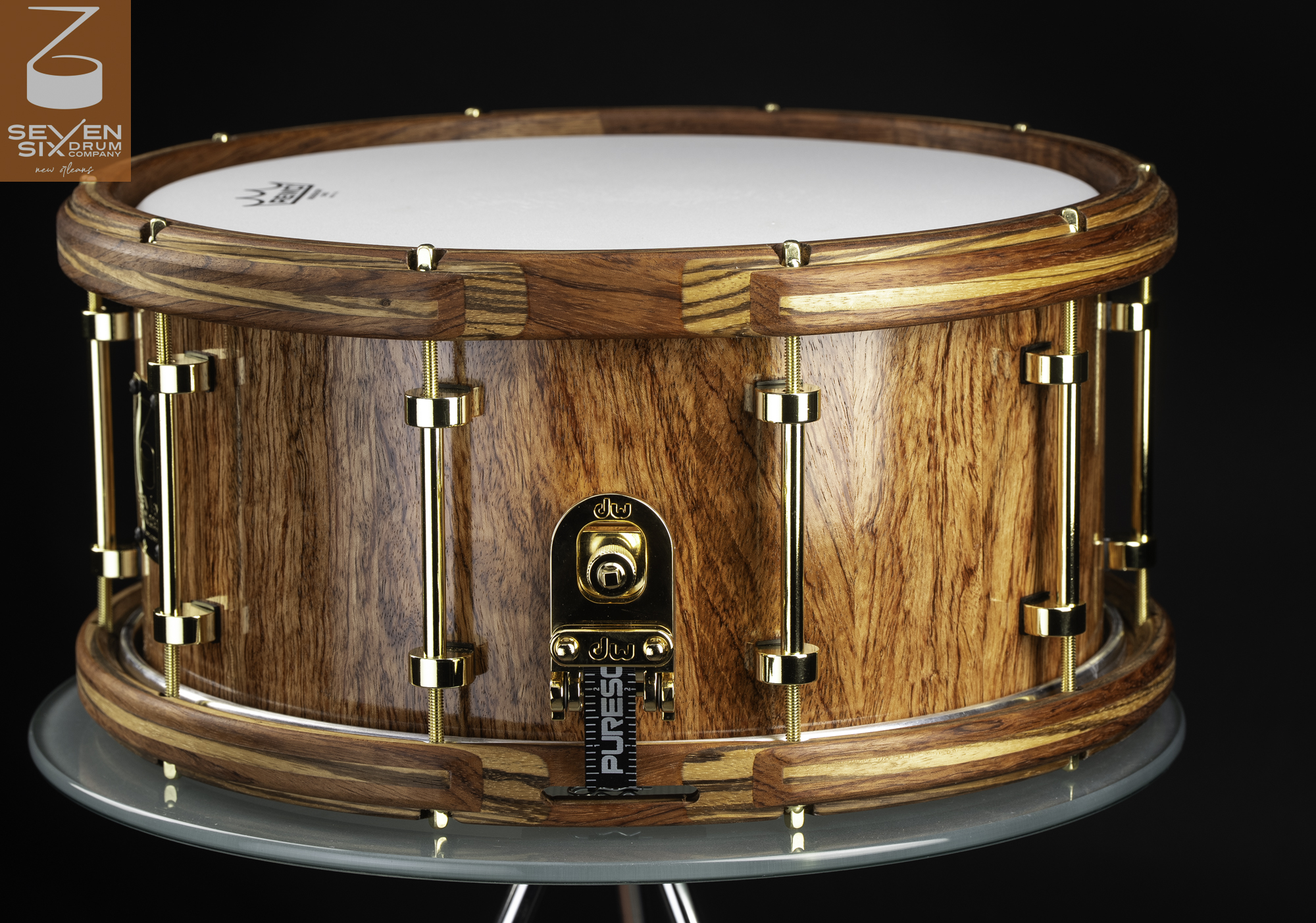 Glossy natural wood snare drum featuring segmented wooden hoops, gold tube lugs, and a gold throw-off.