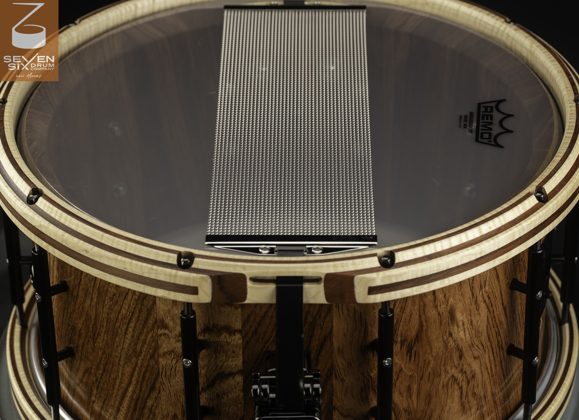 Bottom view of a natural-grain wood snare drum featuring striped wood hoops, black hardware, and wide metal snare wires.