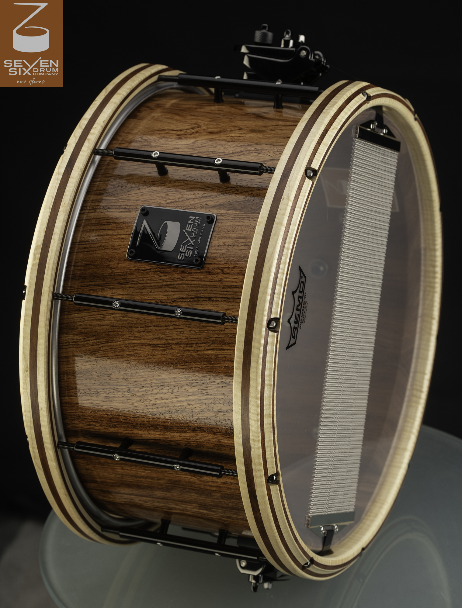 Glossy natural wood snare drum with striped wooden hoops, black tube lugs, and a black throw-off.