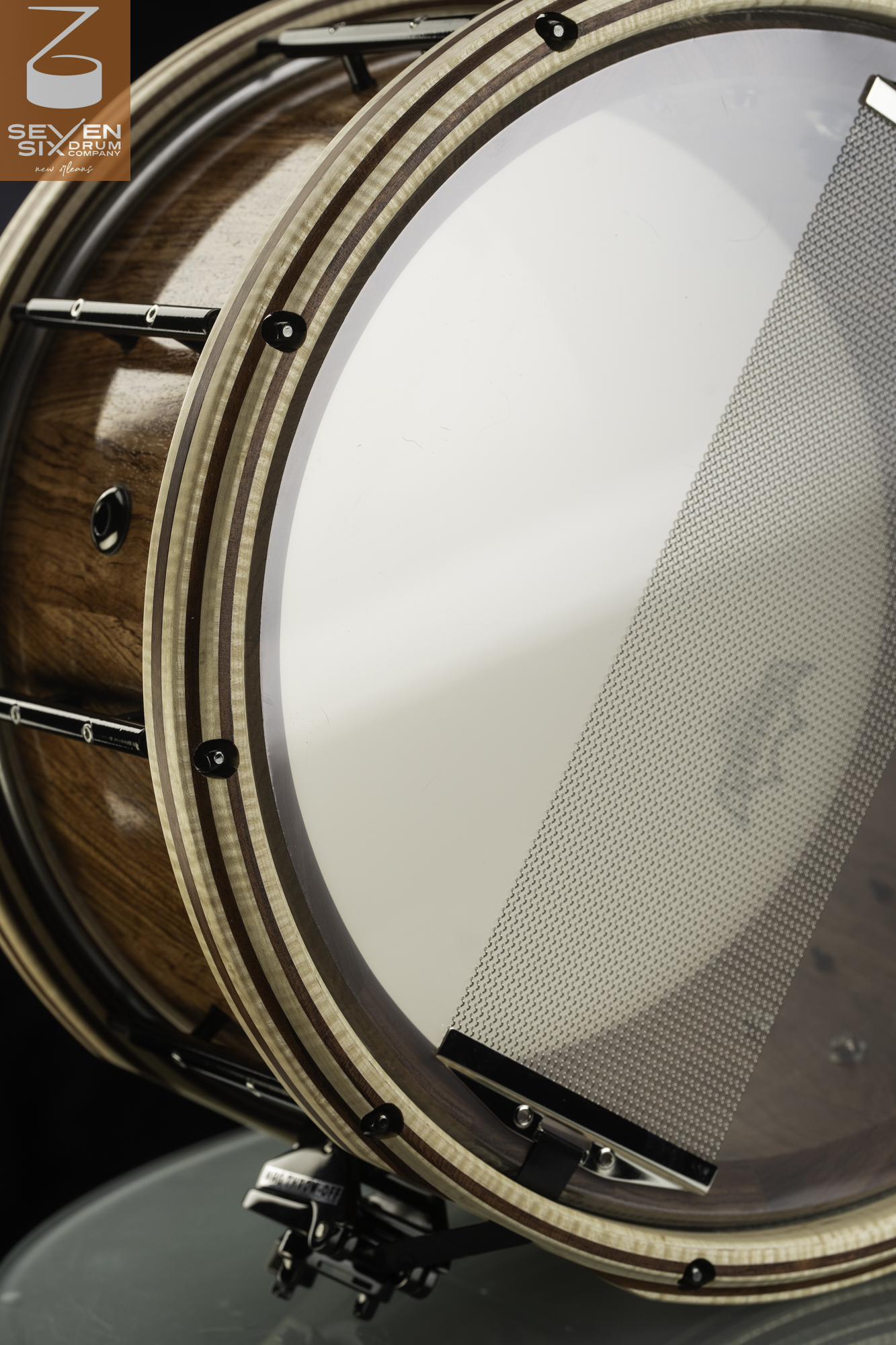 Glossy natural wood snare drum with striped wooden hoops, black hardware, and wide snare wires across a clear resonant head.