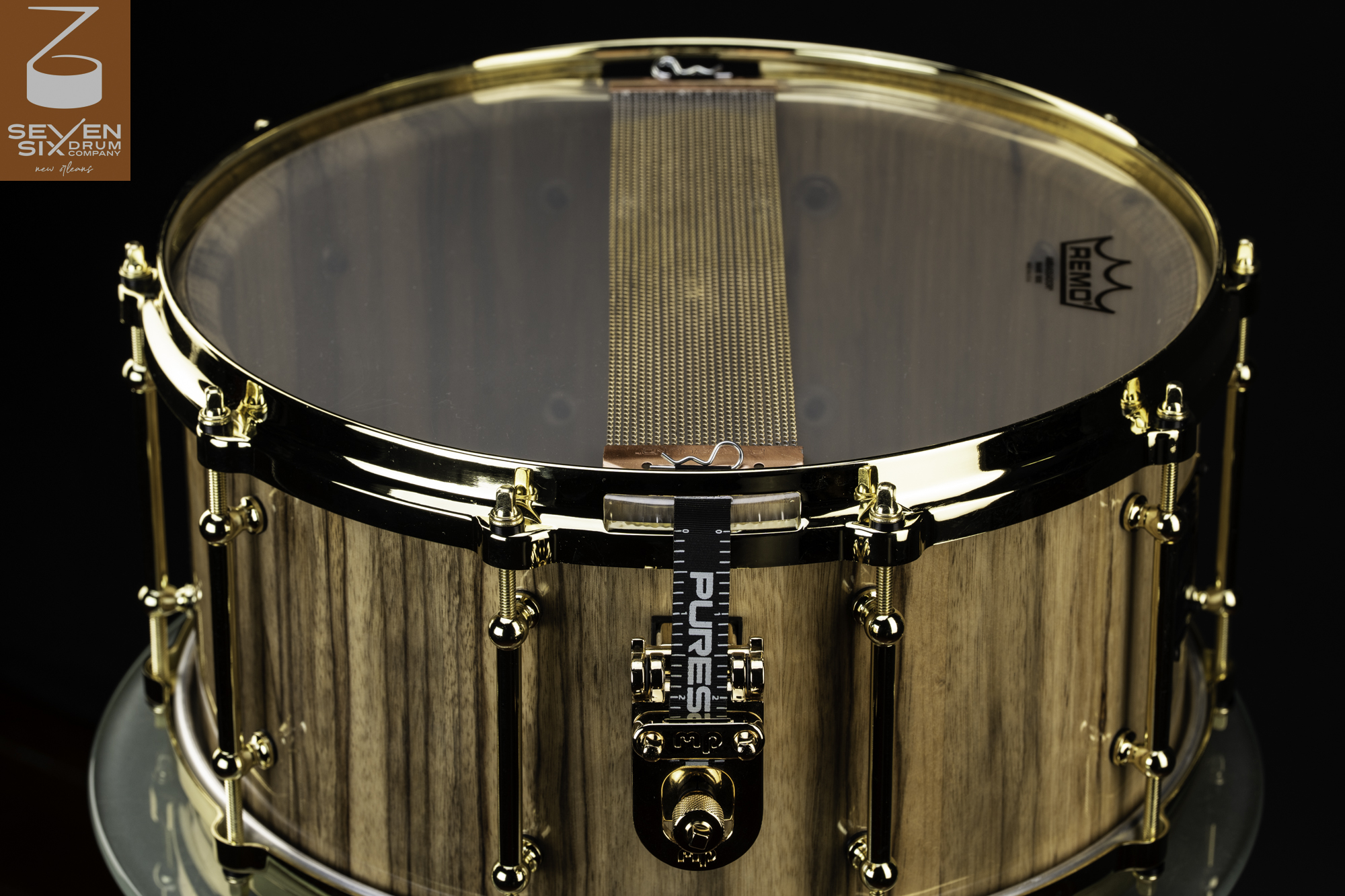 Handmade natural wood snare drum with polished gold hardware, tube lugs, and brass snare wires across a clear bottom head.