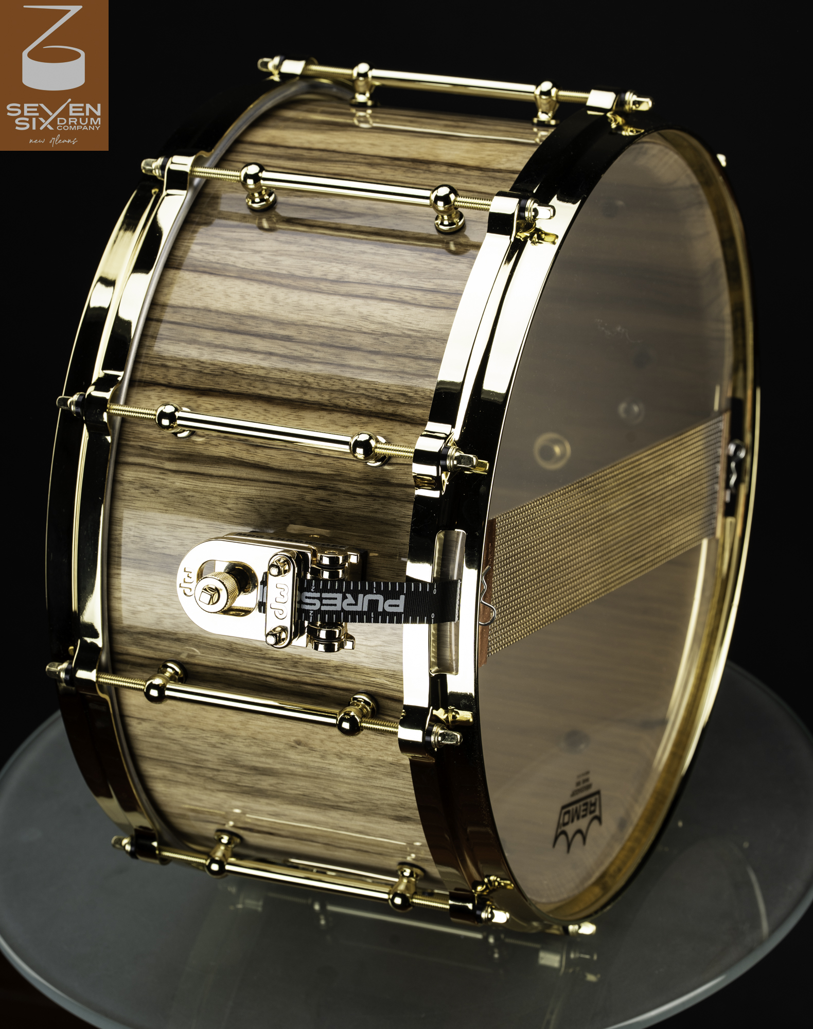 Snare drum crafted from natural wood with a gloss finish, fitted with polished gold hoops, tube lugs, and snare wires.