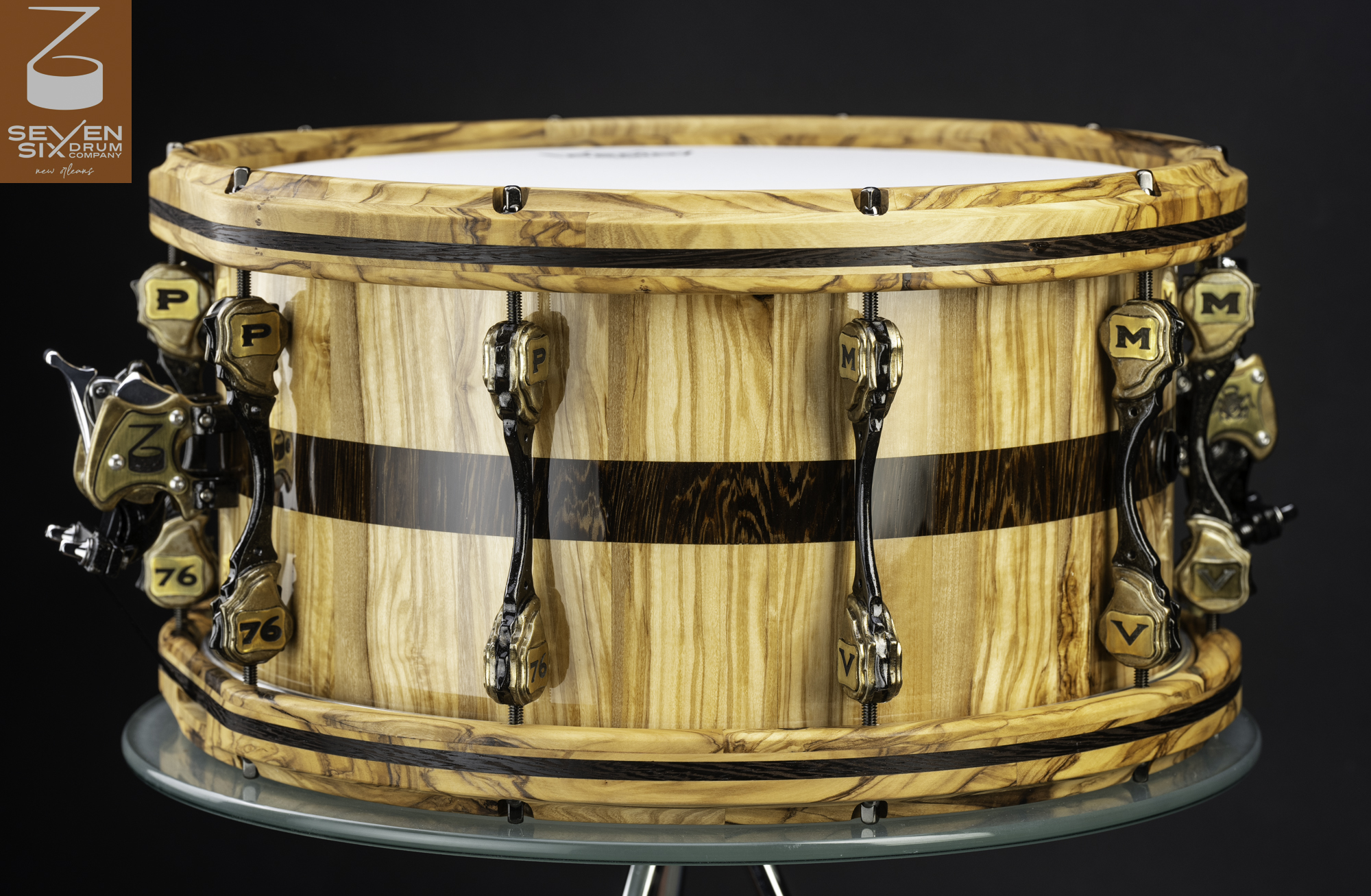 Glossy wood snare drum with a dark center stripe, matching wooden hoops, and custom antique brass and black lugs.