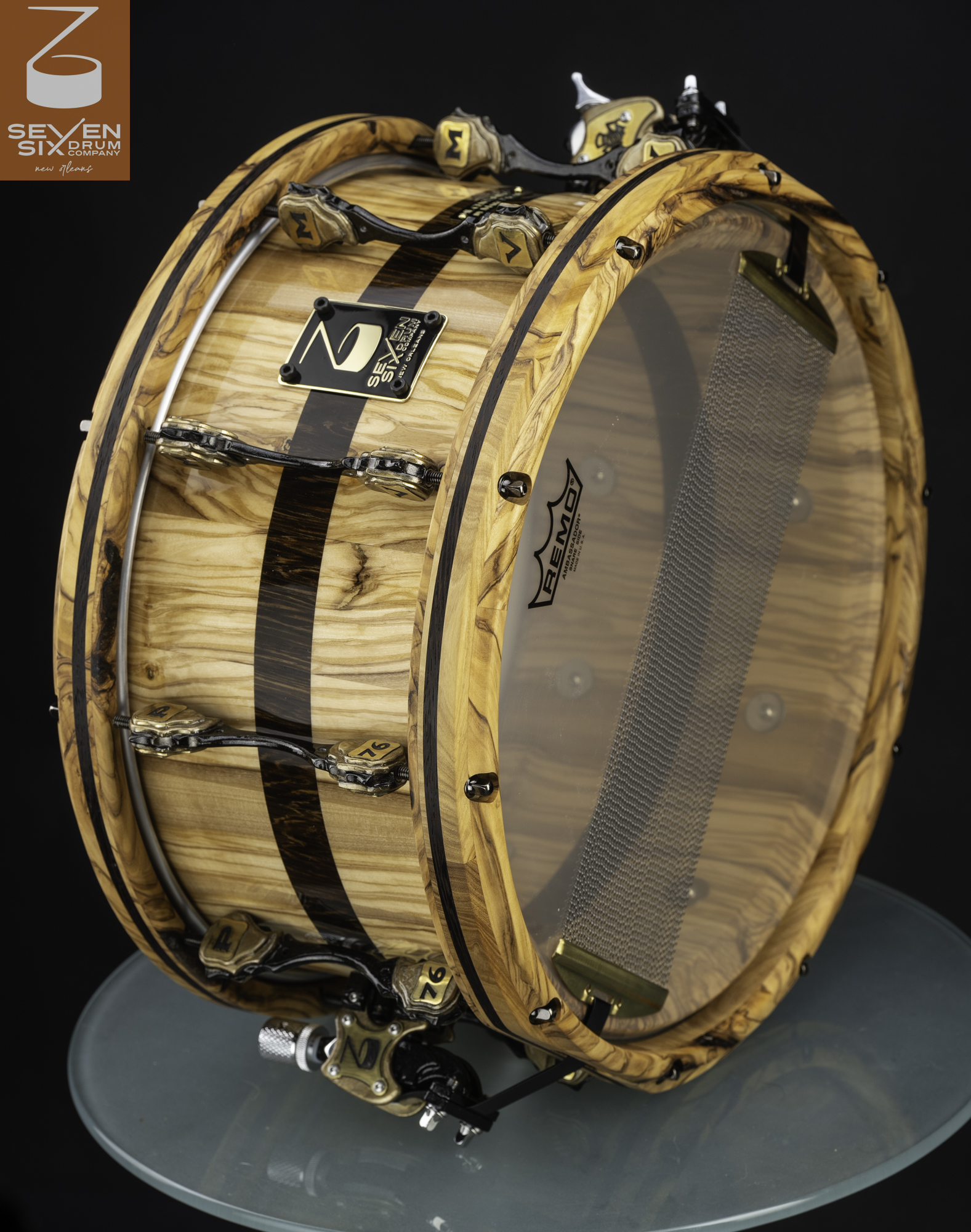 Glossy olive wood snare drum with a dark center inlay, matching wooden hoops, and custom black and brass hardware.