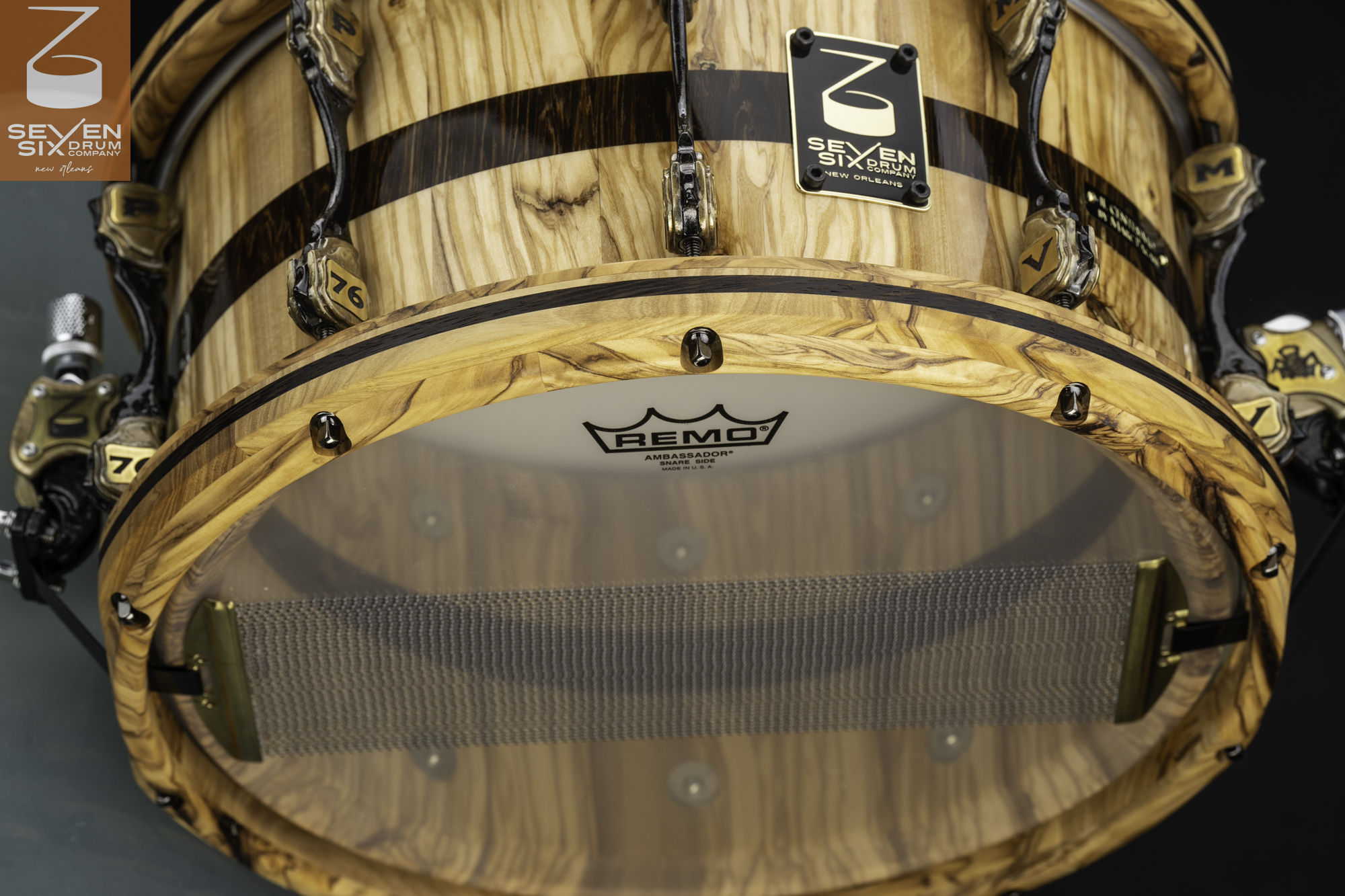Glossy olive wood snare drum with matching wood hoops, dark inlay, custom antique brass lugs, and wide snare wires.